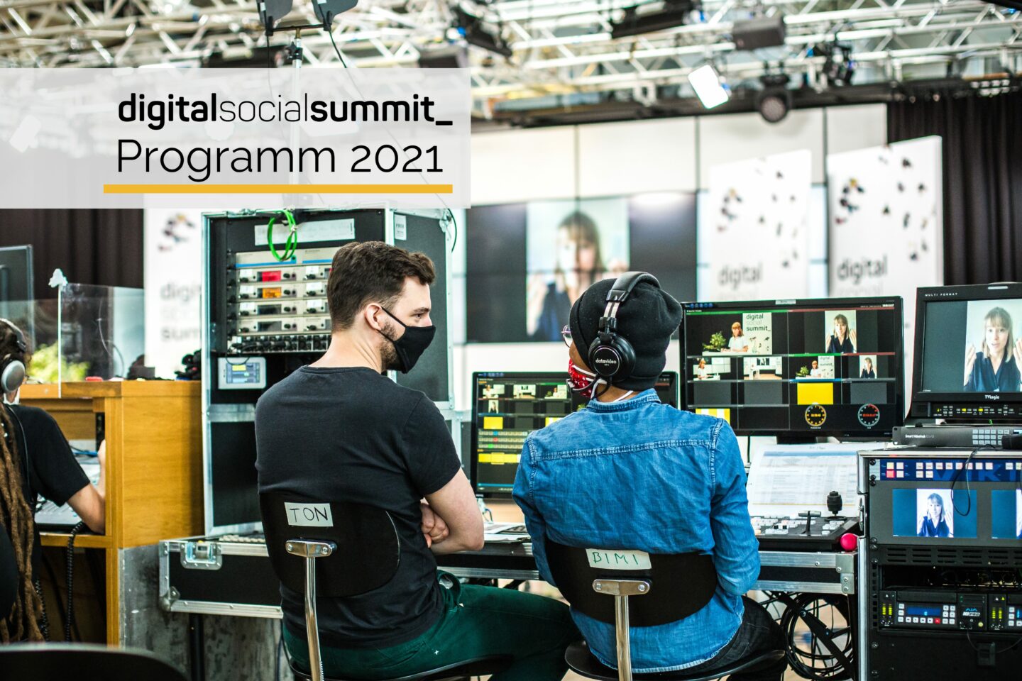 Es ist eine Studioszene zu sehen. Zwei Techniker sitzen nebeneinander vor Monitoren und Schaltpulten. Beide tragen eine Maske und scheinen in ein Gespräch vertieft zu sein. Im Hintergrund ist das Studioset des Digital Social Summits 2020 zu sehen. Oben rechts steht das Logo des Digital Social Summits und Programm 2021.